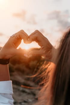 A woman's hands form a heart shape against a golden sunset.