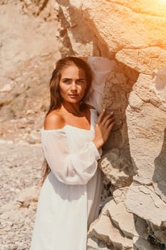 A woman in a white dress stands against a rocky cliff, sun shining on her. She is looking directly at the camera.