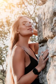 A woman with long hair and a black tank top leans against a rocky cliff, sunlight shining on her face.