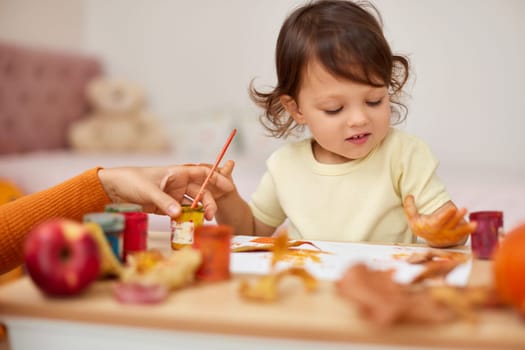 smiling little child girl painting her hands and autumn leaves. baby girl drawing hands in yellow paint