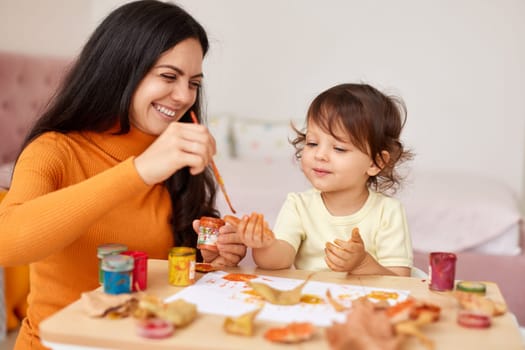 smiling little girl and mother spending good time painting hands and autumn yellow leaves together