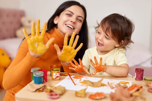 happy little child girl and mother spending good time painting hands and autumn yellow leaves together. smiling mom and baby showing hands in yellow paint