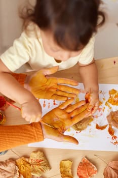 little child girl and mother painting hands and autumn leaves together. mom and baby showing hands in yellow paint. top view