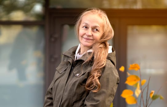 An elderly 60 beautiful woman with a kind smile is walking outdoors on a sunny autumn day, a happy old lady, a grandmother takes care of her appearance, natural beauty with wrinkles portrait closeup.