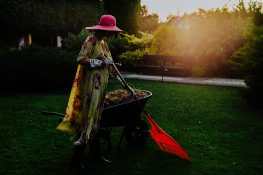A woman dressed in a floral summer dress and wide-brimmed sun hat is seen tending to a garden during sunset. She walks with a wheelbarrow across a beautifully manicured lawn, surrounded by tall trees and lush greenery