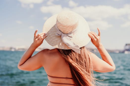 A woman in a swimsuit sits with her back holding a hat, looks at the ocean, sunny day, relaxes