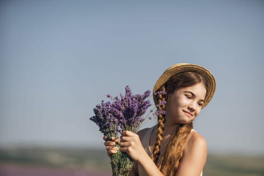 girl is holding a bunch of lavender purple flowers in her hands and wearing a straw hat. She is smiling and she is enjoying herself. The scene is set in a field of lavender, which adds to the peaceful