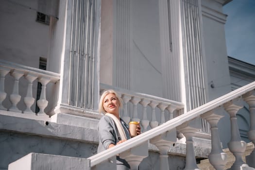 A woman is standing on a white staircase with a cup in her hand. She is smiling and looking up at the sky