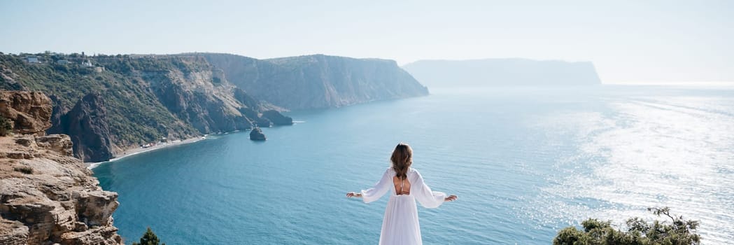 A woman stands on a cliff overlooking the ocean in a white dress. She is looking out at the water and she is in a state of peace and serenity. Concept of calm and tranquility