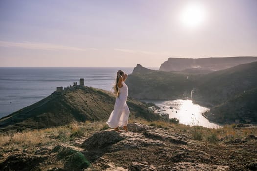 woman stands on a rocky hill overlooking a body of water. She is wearing a white dress and she is in a state of joy or celebration. Concept of freedom and happiness