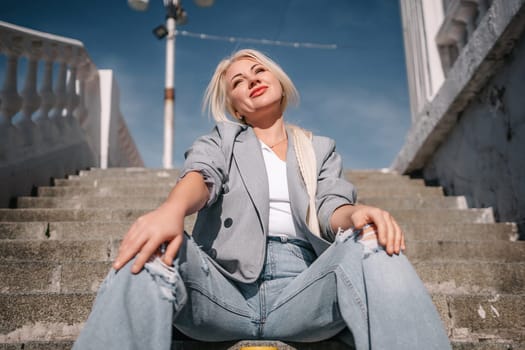A woman in a gray jacket and blue jeans sits on a set of stairs. She is smiling and she is enjoying herself
