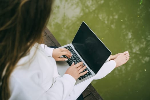 A woman is sitting on a dock with a laptop open in front of her. She is typing on the keyboard and she is focused on her work. Concept of productivity and concentration