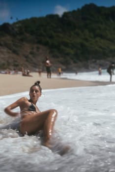 A carefree woman lounges in the surf, surrounded by cheerful beachgoers, embodying summer fun and enjoyment