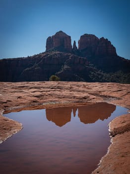 This shot captures the iconic Cathedral Rock in Sedona, Arizona beautifully reflected in a tranquil pool of water. The stark contrast of the red rocks against the blue sky offers a majestic landscape.