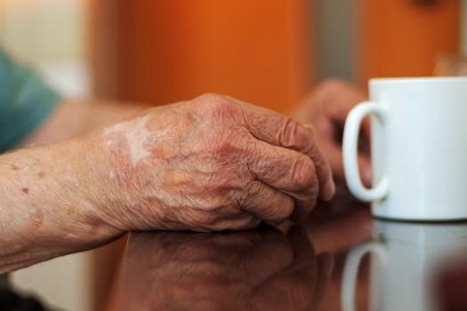 An elderly person hands are gently resting on a table near a white mug.