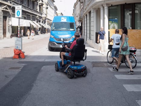 Cremona, Italy August 31st 2024 Senior man driving a mobility scooter crossing the street in the city center with pedestrians and a van in the background