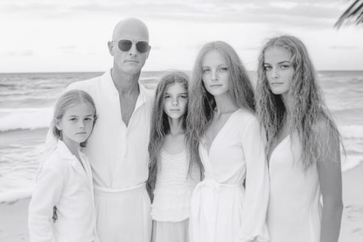 Family wearing white linen outfits is standing together on the beach, posing for a portrait with the ocean in the background