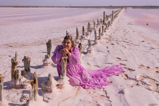 A woman in a pink dress kneels beside weathered wooden posts in a vast expanse of pink salt flats.