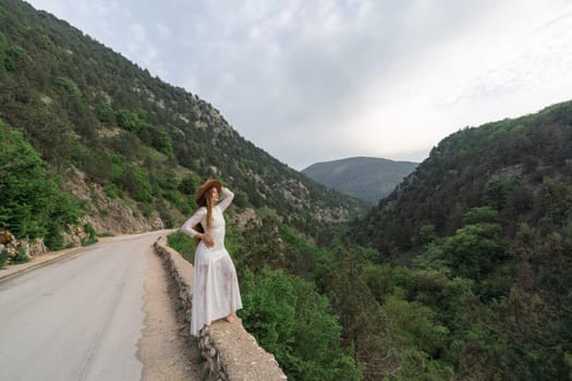 A woman in a white dress stands on a road overlooking a mountain range. The scene is serene and peaceful, with the woman posing for a photo