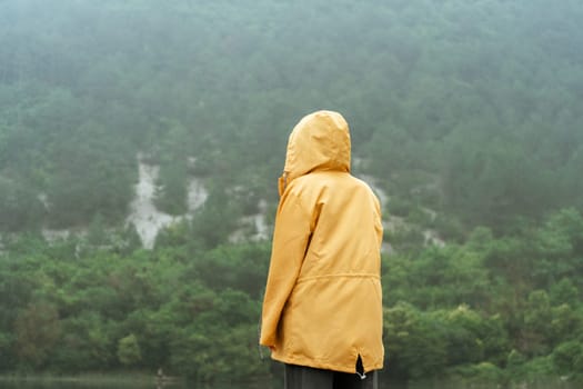 A person is standing in the woods wearing a yellow jacket. The sky is cloudy and the trees are green