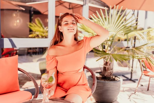 A woman in an orange dress sits at a table with a drink in a glass. The scene is set in a restaurant or cafe, with a potted plant nearby. The woman is enjoying her time, and the atmosphere is relaxed