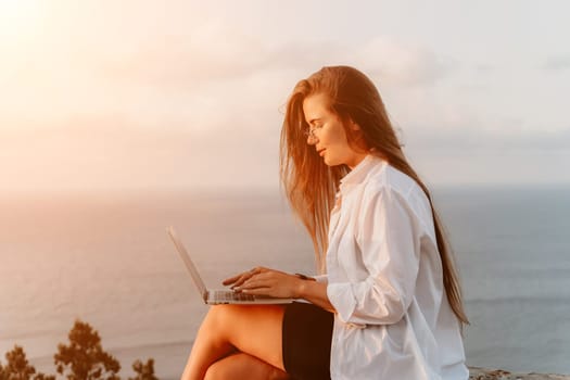 A woman sits on a cliff, typing on a laptop, with the ocean and sunset behind her.
