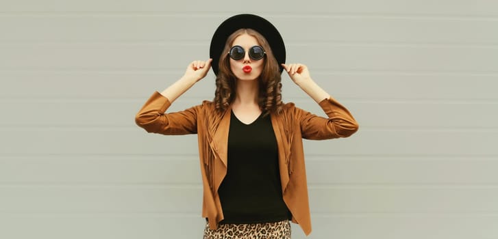 Portrait of beautiful young woman blowing a kiss, stylish girl in black round hat posing on gray background