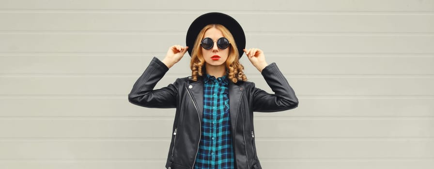 Beautiful stylish young woman posing in black round hat, leather jacket, glasses on city street on gray background