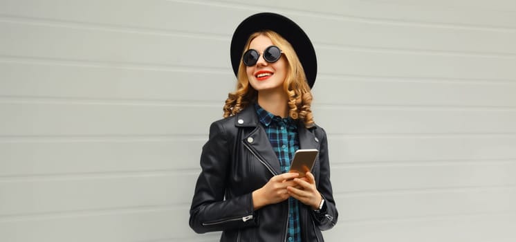Stylish happy smiling modern young woman with phone, girl holding smartphone in black hat, jacket, looking away on gray background