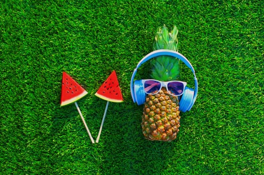 Summer pineapple with headphones listening to music and lollipop or ice cream watermelon on grass