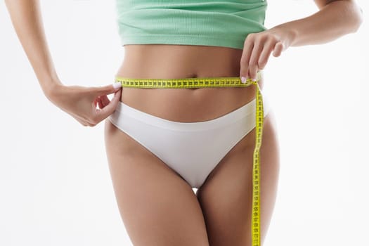A woman measures her waist with a yellow tape measure, she is wearing a green top and white underwear. concept of excess weight, and losing weight