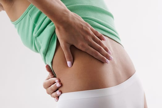 A close-up of a woman's back, showing her hand resting on her lower back. She is wearing a light green tank top and white underwear. indicating discomfort or pain in the lower back area.