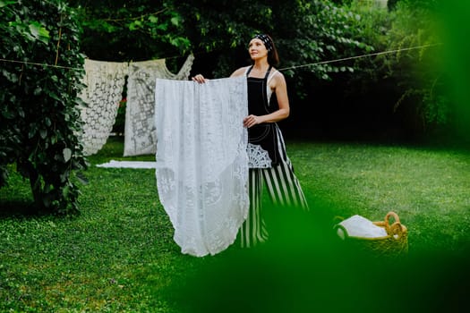 Woman stands in a lush green garden, hanging intricate lace fabric on a clothesline.