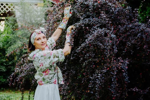 Woman dressed in a floral gardening outfit is pruning a large shrub in a lush garden.