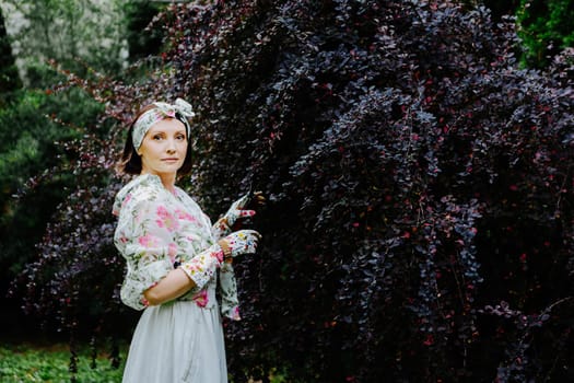 Woman dressed in a floral gardening outfit is pruning a large shrub in a lush garden.