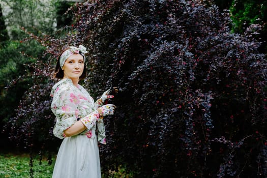 Woman dressed in a floral gardening outfit is pruning a large shrub in a lush garden.