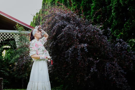 Woman dressed in a floral gardening outfit is pruning a large shrub in a lush garden.
