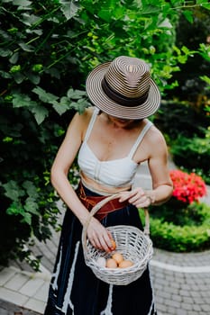 A woman in a stylish summer outfit and a striped sun hat is collecting eggs in a wicker basket in a lush garden.