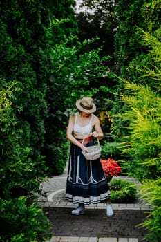 A woman in a stylish summer outfit and a striped sun hat is collecting eggs in a wicker basket in a lush garden.