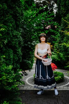 A woman in a stylish summer outfit and a striped sun hat is collecting eggs in a wicker basket in a lush garden.