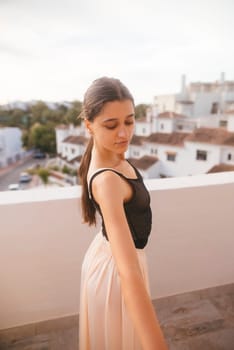 A young dancer strikes a graceful pose on a balcony with a scenic backdrop of picturesque white buildings