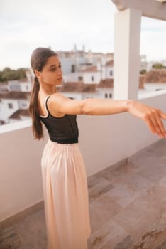 A young woman wearing a flowing skirt strikes a graceful pose on a balcony that overlooks a vibrant landscape