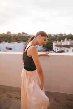 A graceful woman wearing a flowing skirt elegantly poses on a beautiful balcony during a serene sunset scene