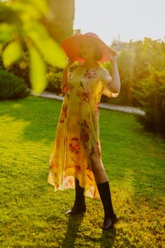 A woman wearing a floral dress and a large pink sun hat stands in a lush garden during golden hour. She holds the brim of her hat, casting a soft shadow over her face as the sunlight illuminates the scene.