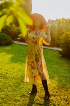 A woman wearing a floral dress and a large pink sun hat stands in a lush garden during golden hour. She holds the brim of her hat, casting a soft shadow over her face as the sunlight illuminates the scene.