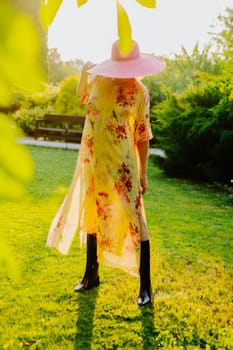 A woman wearing a floral dress and a large pink sun hat stands in a lush garden during golden hour. She holds the brim of her hat, casting a soft shadow over her face as the sunlight illuminates the scene.
