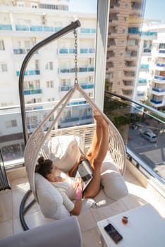 A woman lounges in a hanging chair, enjoying her tablet while relaxing on a peaceful, sunny balcony