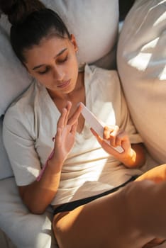A young woman is lounging comfortably on a plush sofa, deeply focused on her smartphone while enjoying relaxation
