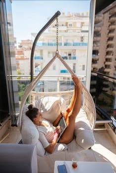 A woman enjoys a moment of relaxation in a stylish hanging chair while working on her laptop on a bright, sunny balcony