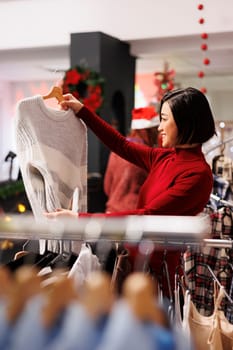 Customer browsing through clothes on racks, looking to buy items as presents for christmas eve at shopping mall. Asian woman buying merchandise on discount, checking fabric.
